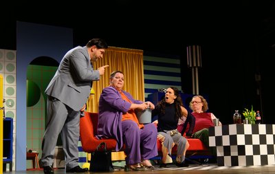 Das Bild zeigt eine emotionale Szene auf einer Theater-Bühne. Vier Personen streiten miteinander. Man sieht die bunte Kulisse mit einer blauen Wand, einem gelben Vorhang und einer gestreiften Lampe