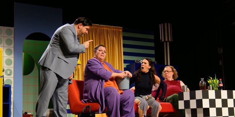 Das Bild zeigt eine emotionale Szene auf einer Theater-Bühne. Vier Personen streiten miteinander. Man sieht die bunte Kulisse mit einer blauen Wand, einem gelben Vorhang und einer gestreiften Lampe