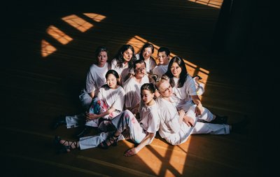Gruppenfoto: 8 Frauen sitzen auf einem Holzfußboden um einen Mann, alle haben weiße Kleidung an