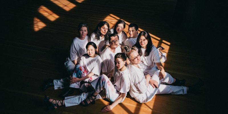 Gruppenfoto: 8 Frauen sitzen auf einem Holzfußboden um einen Mann, alle haben weiße Kleidung an