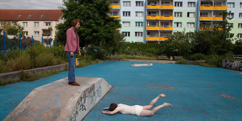 blaufarbige Skaterbahn mit einer auf dem Bauch liegenden Person und einer auf einer Rampe stehenden Person, die auf die Liegende sieht.