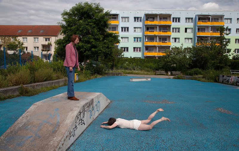 blaufarbige Skaterbahn mit einer auf dem Bauch liegenden Person und einer auf einer Rampe stehenden Person, die auf die Liegende sieht.