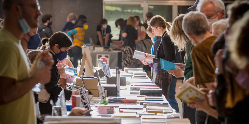 Viele Menschen an einem langen Tisch stehend, auf dem Bücher liegen, die sie ansehen